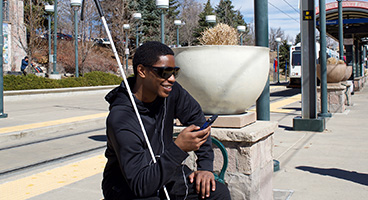 A smiling young blind man sits outside waiting for a train while listening to NFB-NEWSLINE on his phone.